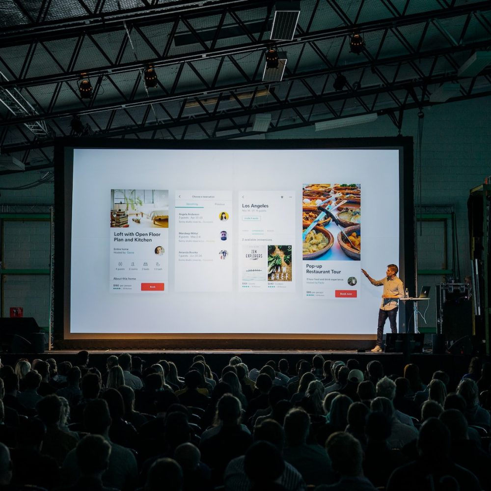 person discussing while standing in front of a large screen in front of people inside dim-lighted room